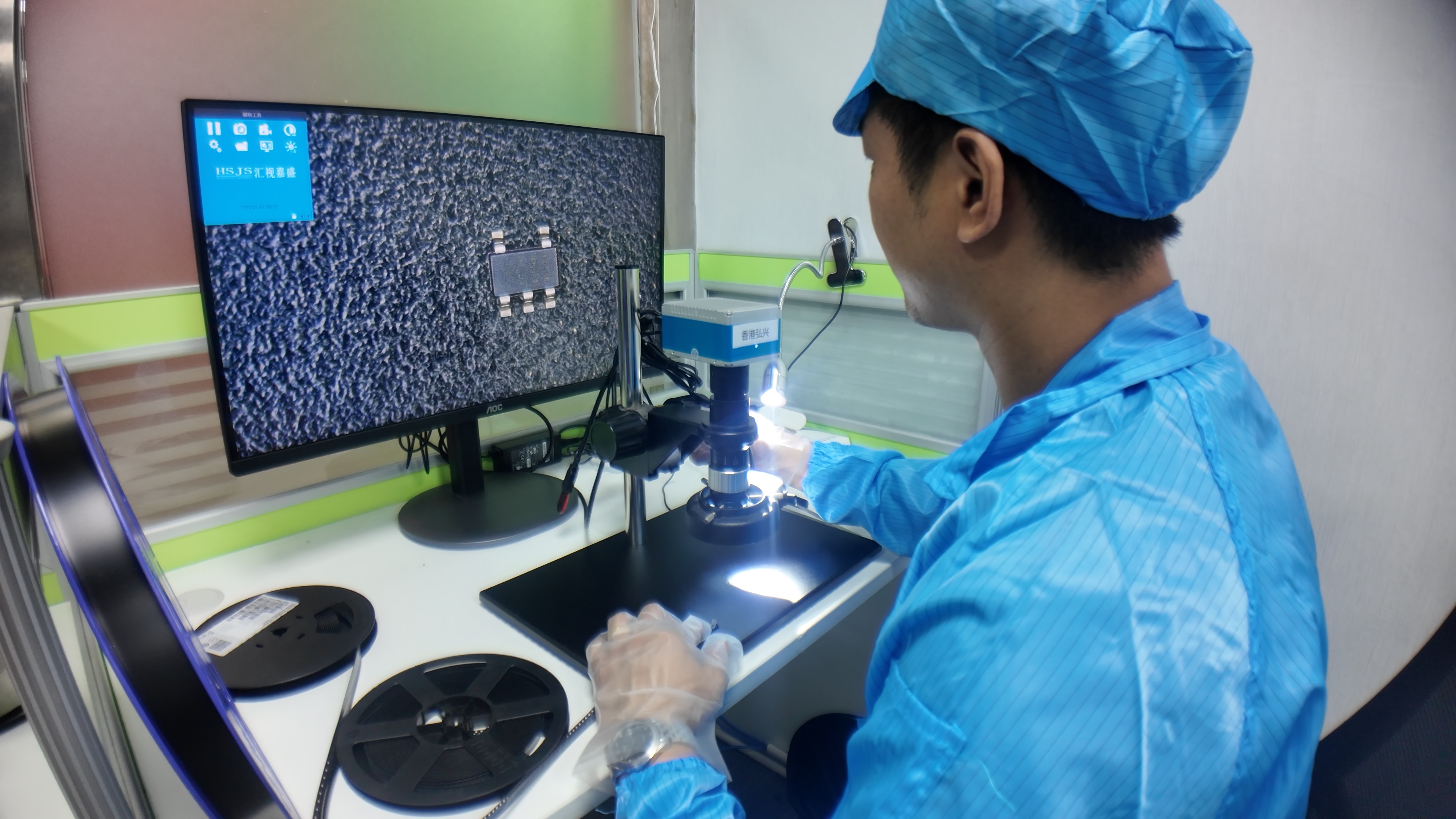 Technician checking a chip on a microscope display, with component reels on the bench